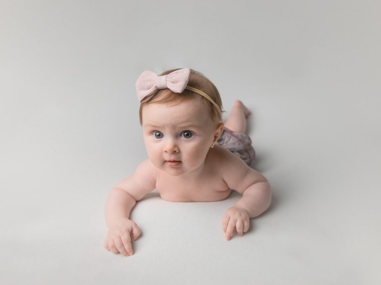 img source='pic.gif' alt=SESIONES BEBES BARCELONA Bebé con una diadema rosa, recostado sobre una superficie blanca.